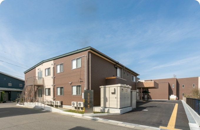 介護施設の建物外観。レンガ造りの2階建て施設で、駐車場と玄関アプローチが見える。