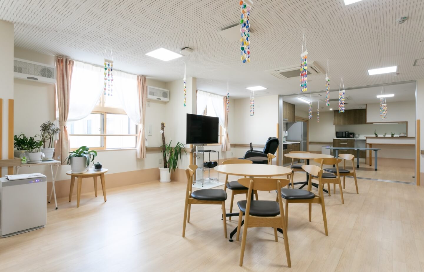 木製家具と装飾が配置された明るい共用スペース。テレビや観葉植物、つるし飾りがあり、入居者の交流空間として利用されている。