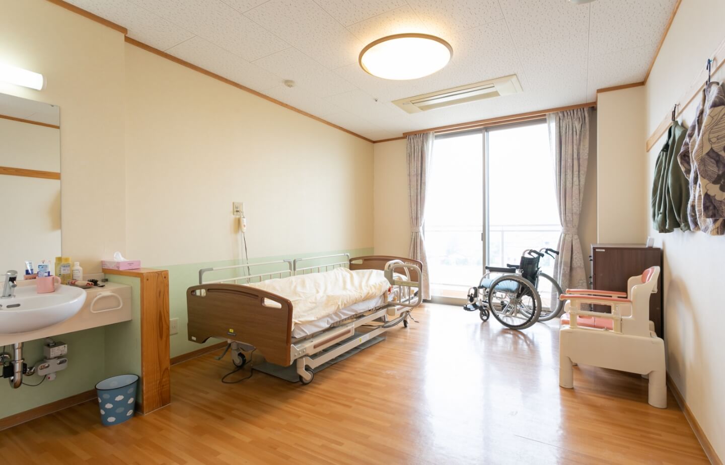 介護ベッド、車椅子、洗面台を備えた居室。明るく清潔な個室空間。