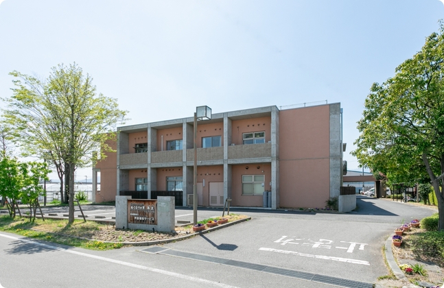 介護施設の建物外観。茶系3階建て、駐車場完備、緑に囲まれた立地