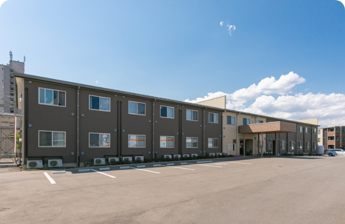 介護施設の建物外観。駐車場からの全景。ダークブラウンと淡色の外壁。